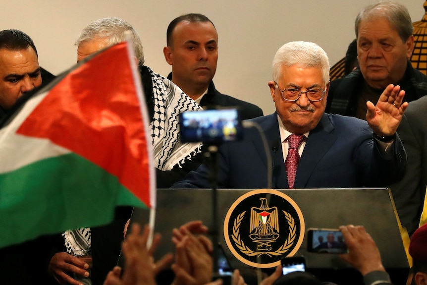 Mahmoud Abbas waves at supporters from a podium. A row of men stand behind him.