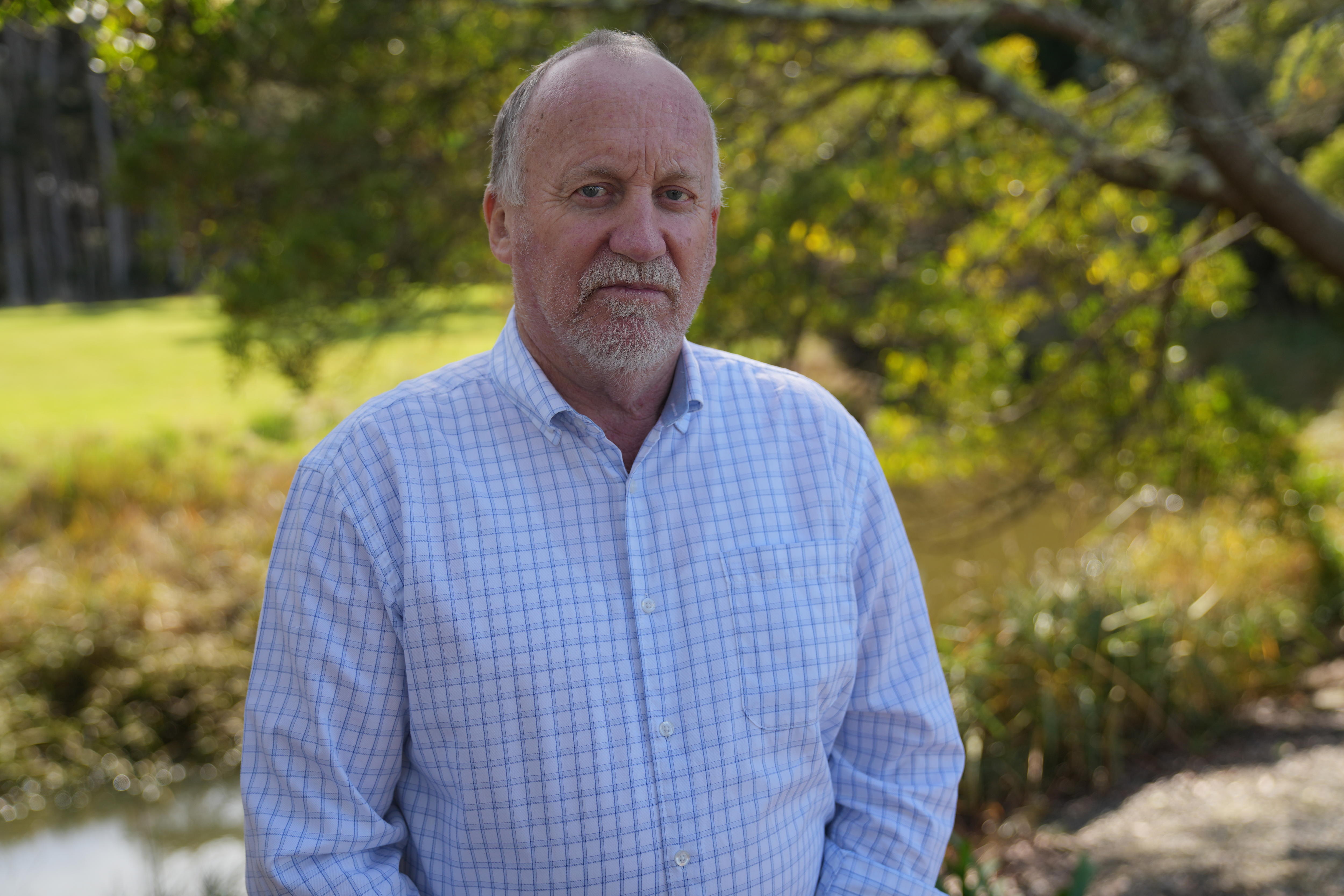 He wears a collared shirt and stares down the camera. A creek and tree are behind him.