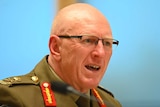 A bald man with glasses and a military uniform speaks with a bright blue background.