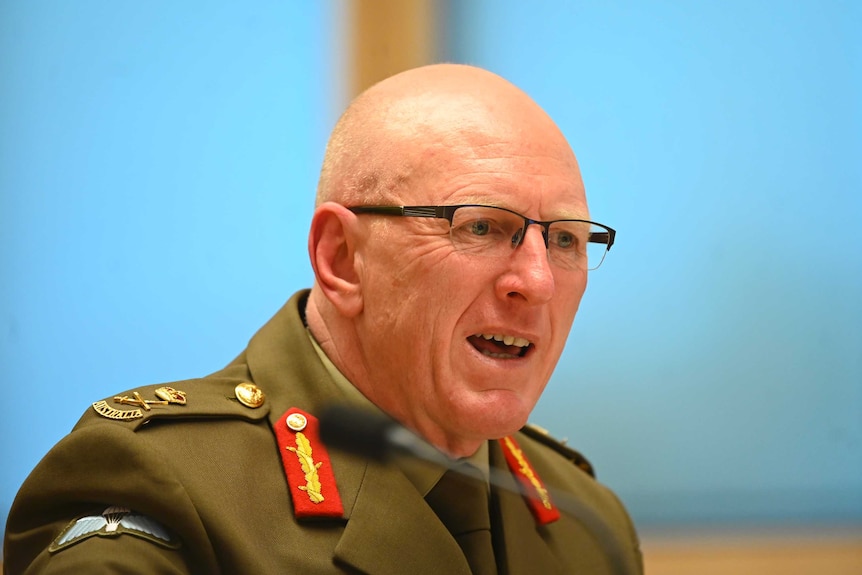 A bald man with glasses and a military uniform speaks with a bright blue background.