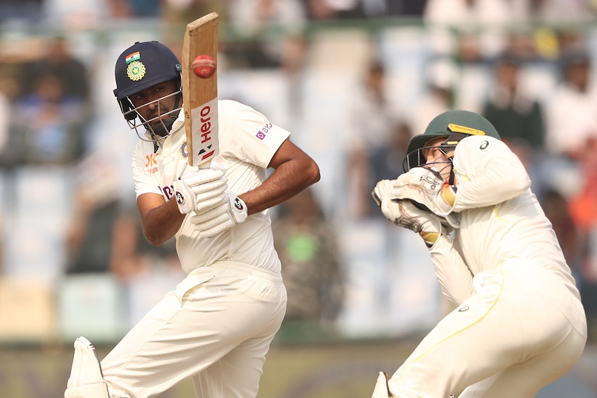 An Indian male Test batter hits a shot as the Australian wicketkeeper looks on.