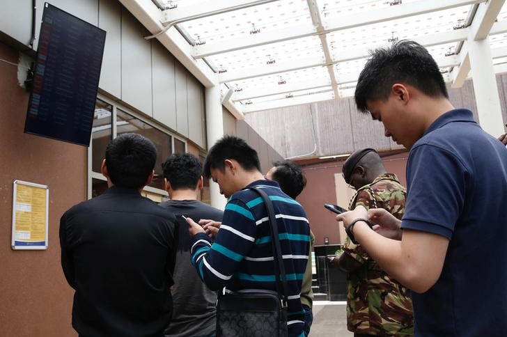 Five travellers look to their phones and at flight information board as Kenyan military officer does the same in background.