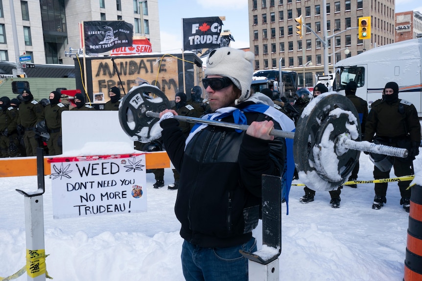 A man with a cigarette in his mouth lifts a barbell wearing a polar bear beanie and sunglasses as riot police approach. 