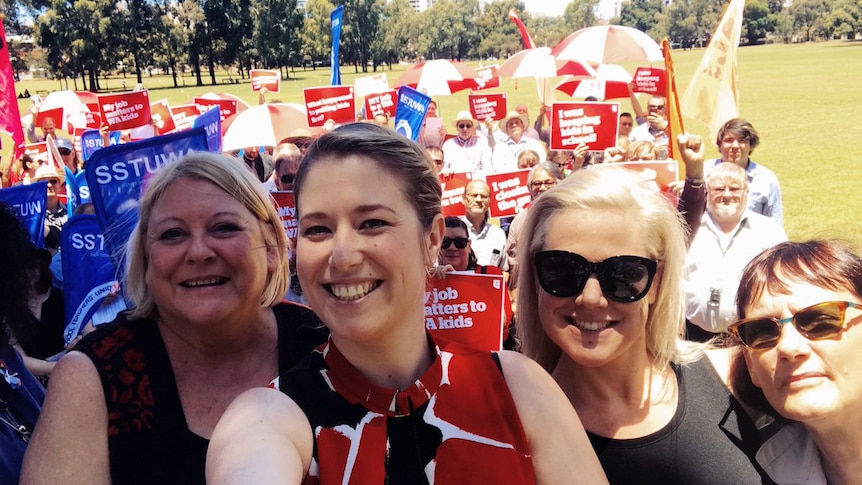 Four women smile in a selfie photo at a union rally with protestors holding banners in the background