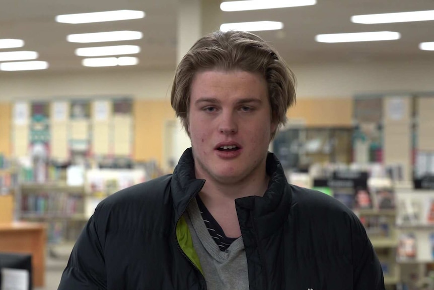 Sam Macrae wearing a black jacket and grey top standing in a library.