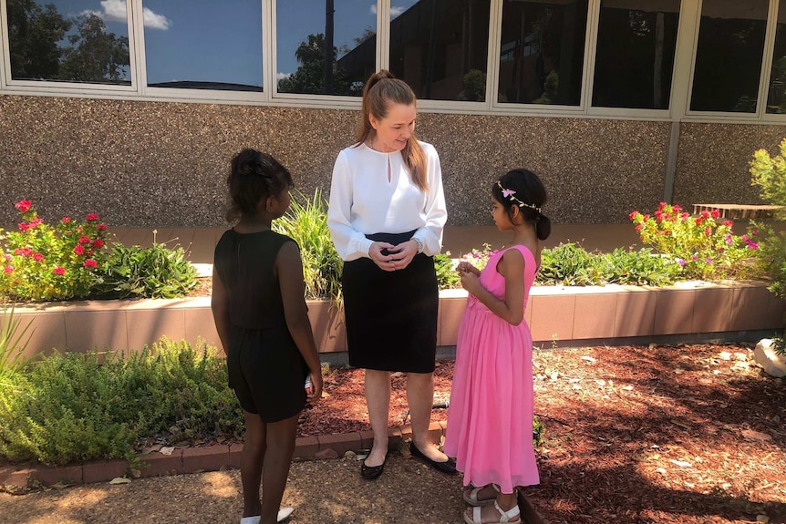 Two young indigenous girls in dresses talk to a woman in business clothing. They are outside a building.