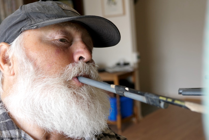 A 67-year-old man holding paint brush in his mouth concentrates on his painting