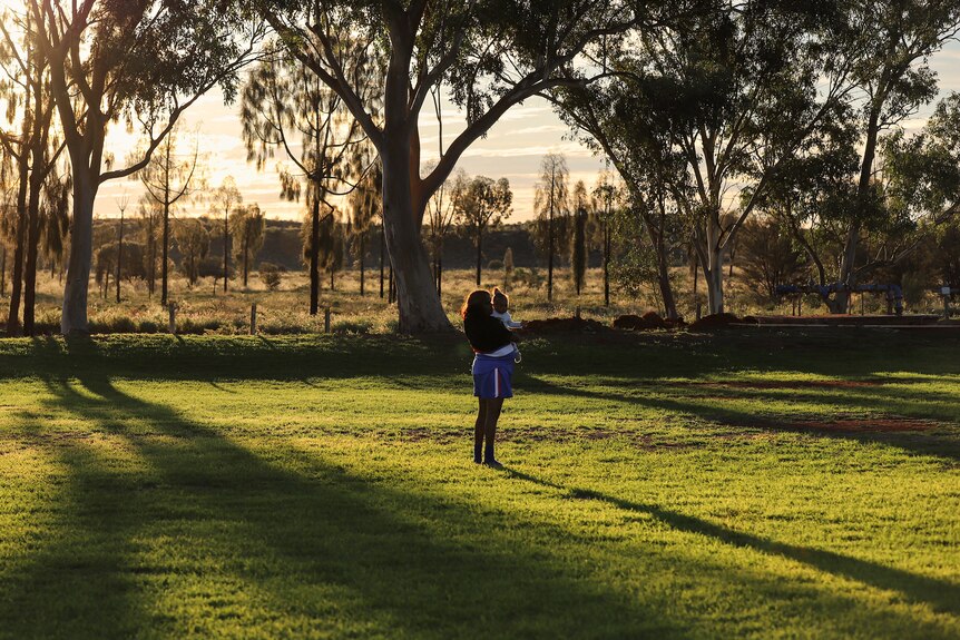 An Aboriginal woman holds a baby standing in the shadows of a late afternoon sun.