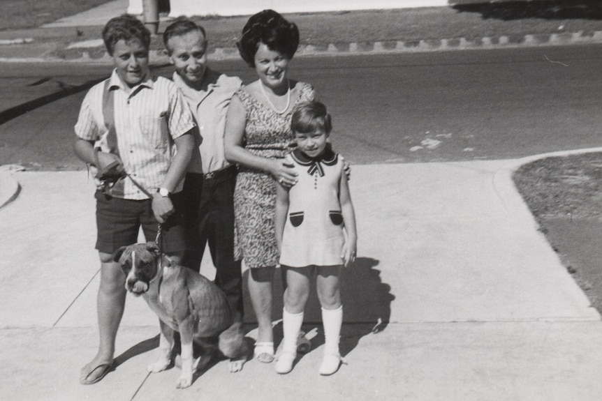 Black and white photo of two parents and two young children, and their dog, in summer clothes, standing on the footpath smiling.