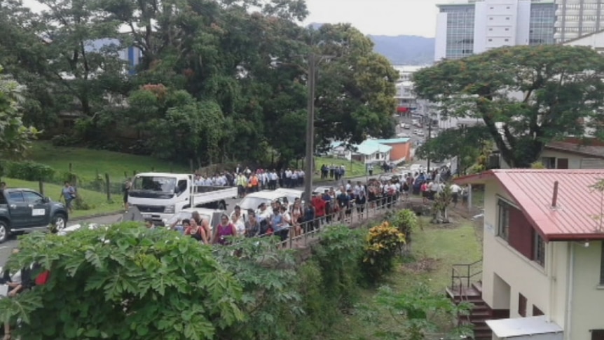 People stand in a line down the street of Fijian city ___.