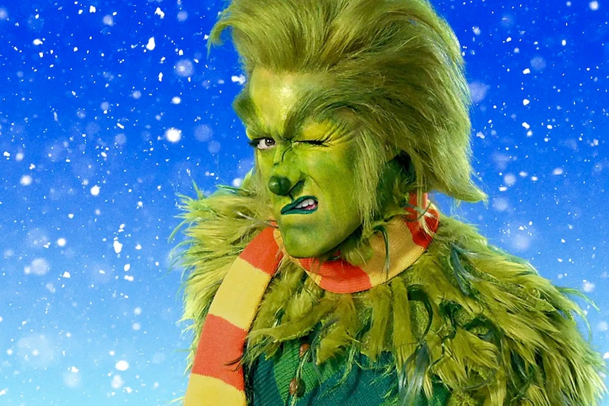 A man in a green hairy Grinch costume winking with snow falling