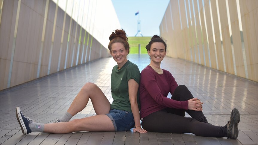 Two women are sitting on the concrete back-to-back, smiling at the camera, with a blurred corridor behind them