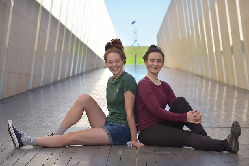 Two women are sitting on the concrete back-to-back, smiling at the camera, with a blurred corridor behind them