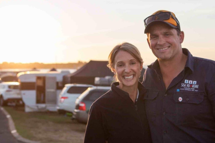 A man and woman smile at the camera with cars and caravans set up behind them