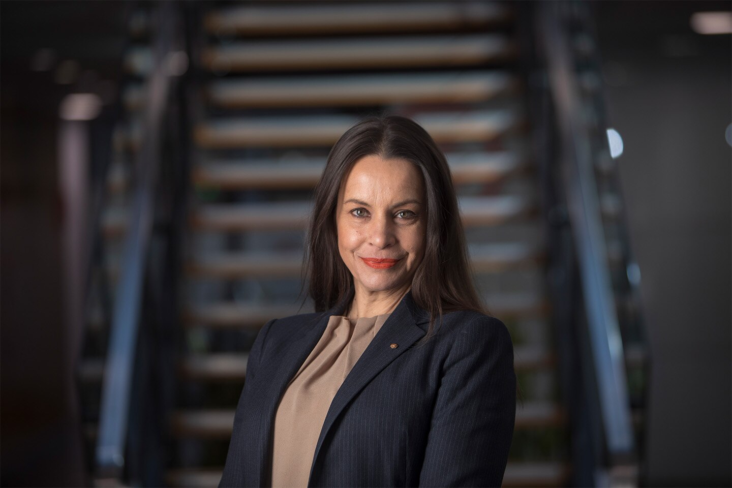 A smiling woman with straight, shoulder-length hair, wearing a suit jacket, standing in front of a blurred staircase.