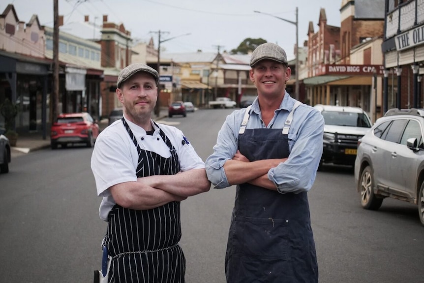 Two smiling men wearing butcher outfits standing on an historic street