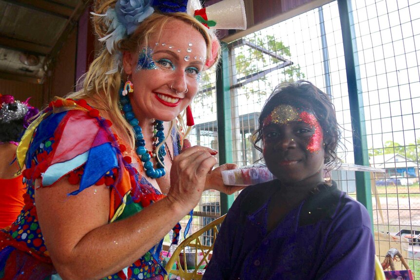 A little girl gets a sparkly Aboriginal flag painted on her face by a woman in a colourful fairy costume.