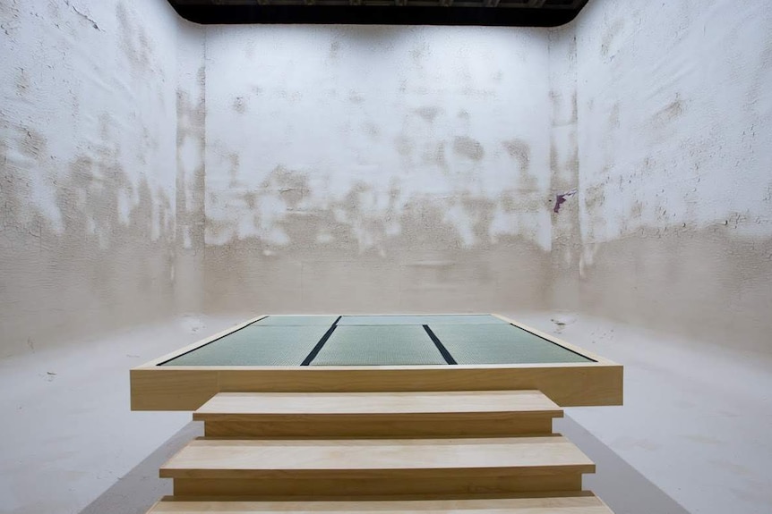 Wooden steps leading to a flat wooden platform in a white, square concrete space.