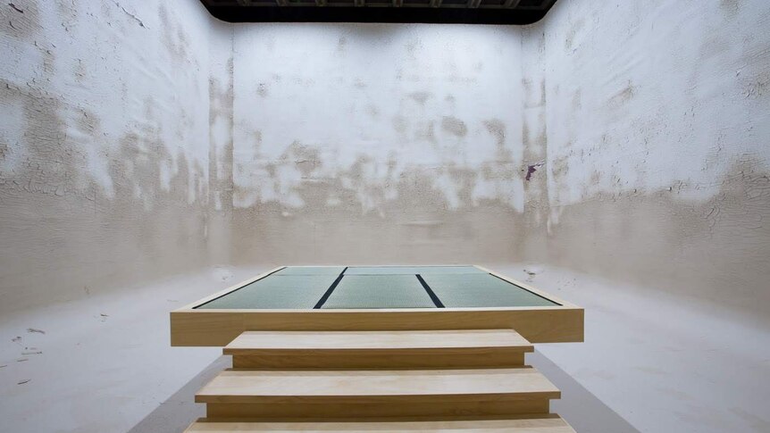 Wooden steps leading to a flat wooden platform in a white, square concrete space.