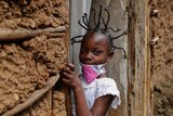 A young girl with her hair tied in spikes looks at the camera as she holds onto a door frame.