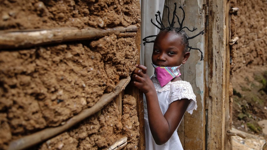 A young girl with her hair tied in spikes looks at the camera as she holds onto a door frame.