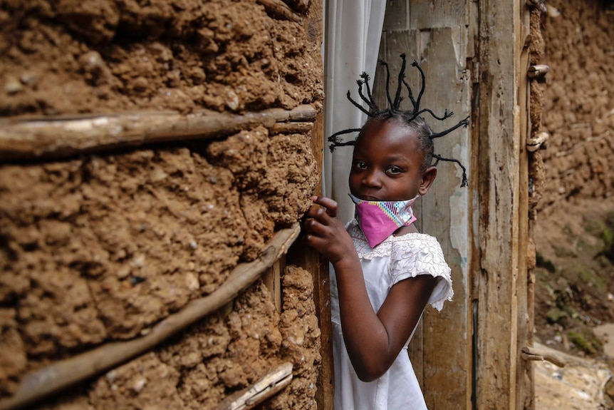 A young girl with her hair tied in spikes looks at the camera as she holds onto a door frame.