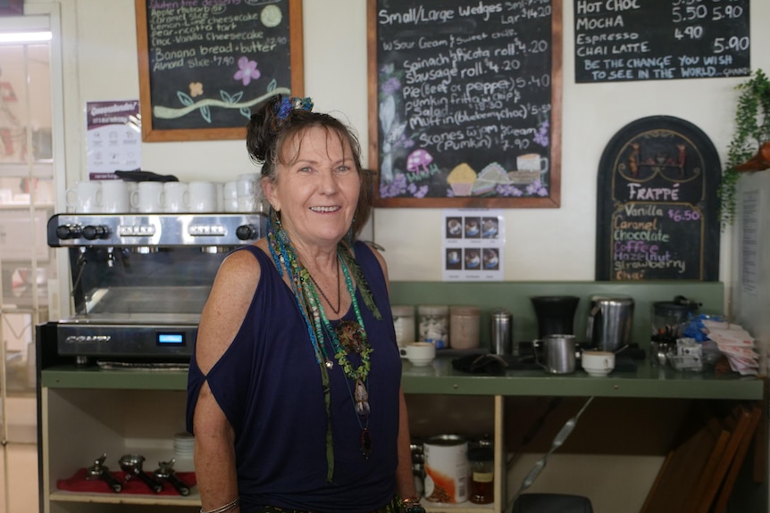 Tess Ford stands in front of a coffee machine and a shelf containing cups and jugs, menus are visible hanging on the wall behind