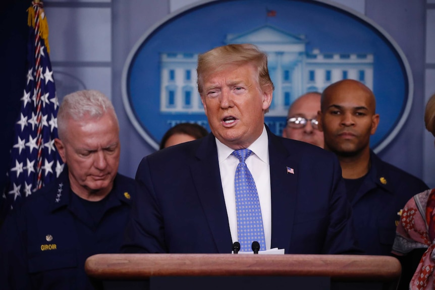 Donald Trump stands at a lectern and speaks. A number of people stand behind him.