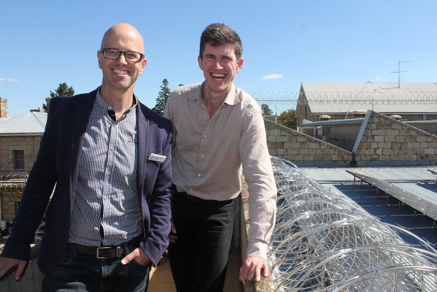 Two men in their thirties stand on a roof with solar panels in the background