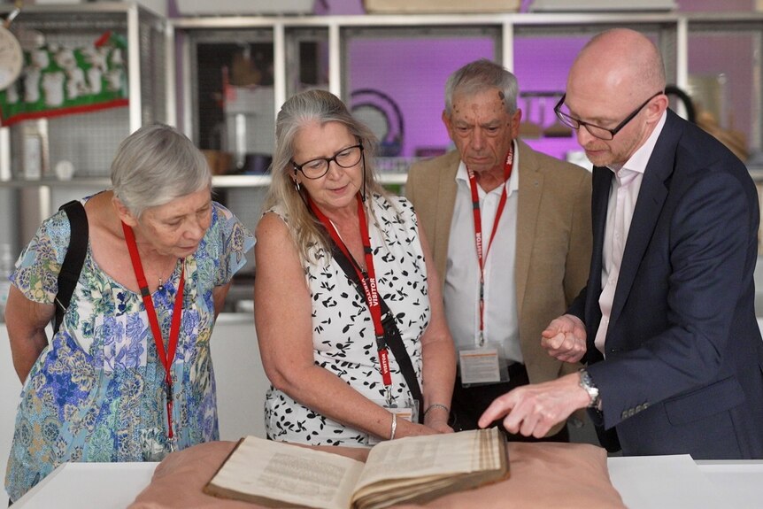Two men and two women, all with grey hair, look down at an old book.