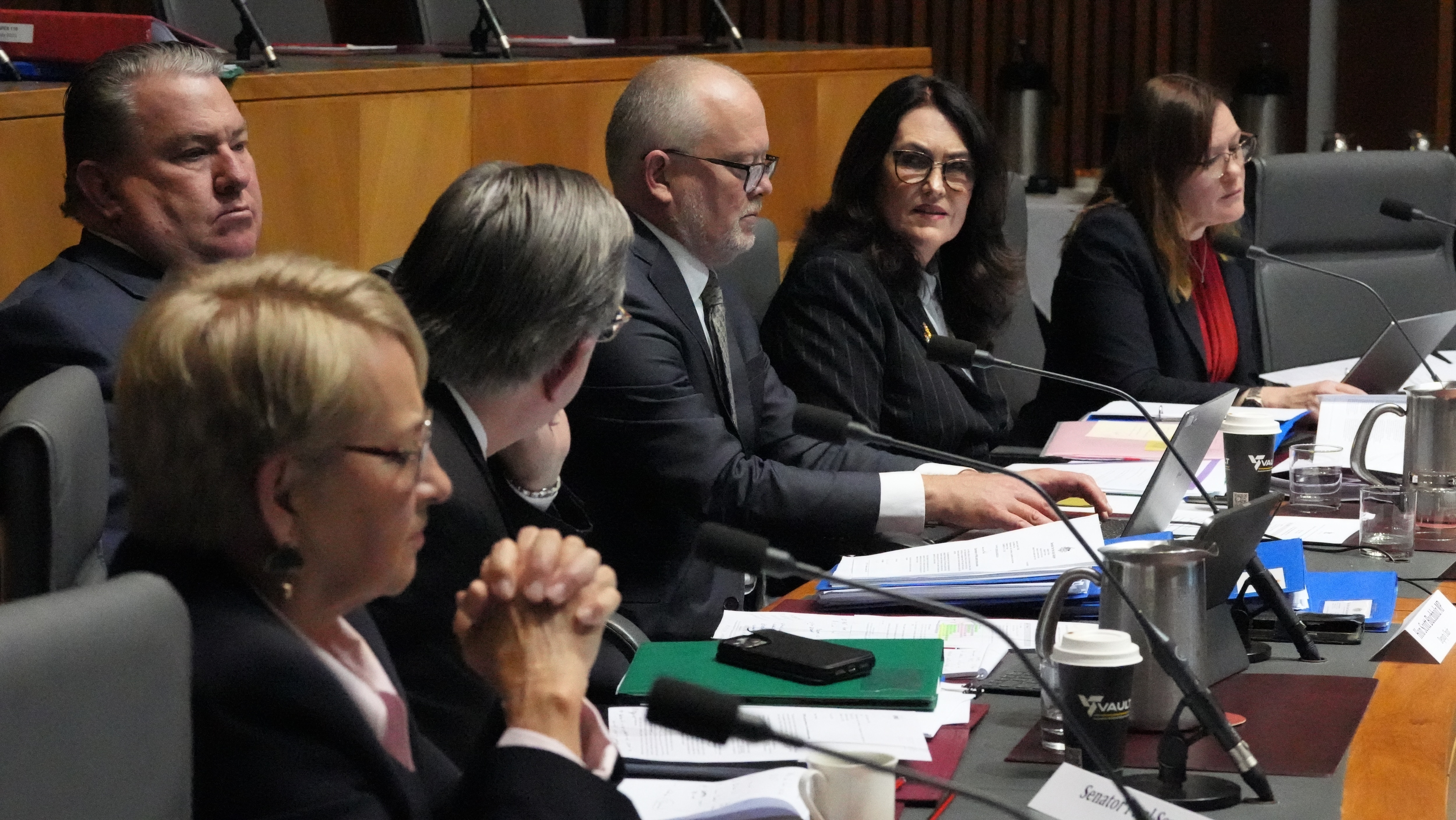 Men and women in dark formal wear sit at a long table in a parliamentary chamber.