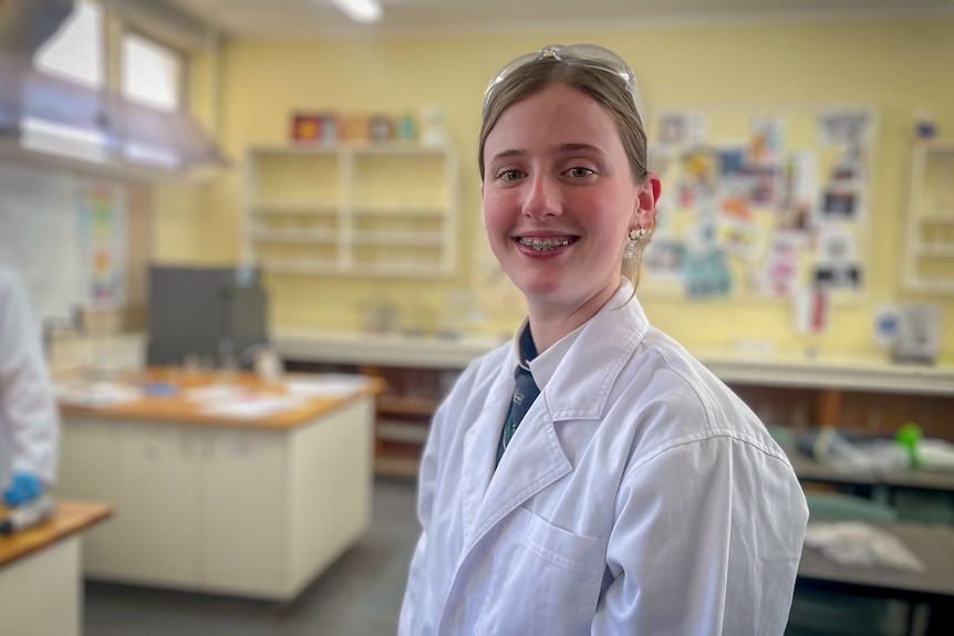 Alanah wears a white lab coat and smiles at the camera during a science class.