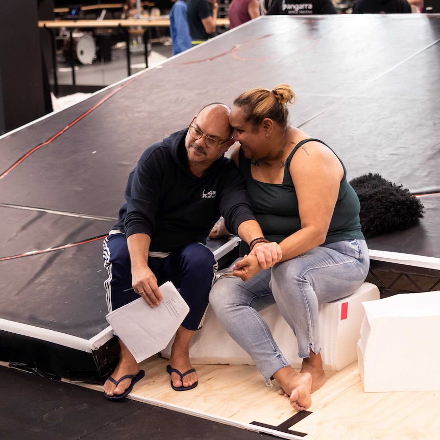 Stephen Page (left) and Elaine Crombie (right) embrace while sitting by an angled stage in a rehearsal room.