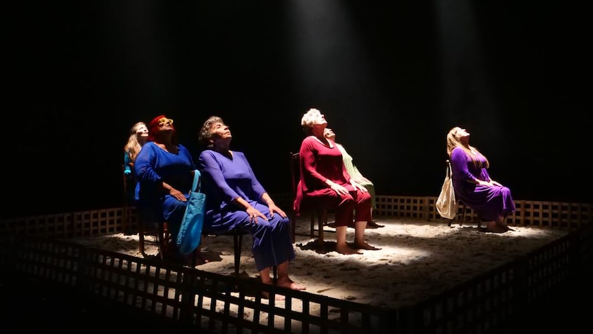 A group of women sit on chairs, looking upwards into bright spotlights on stage.