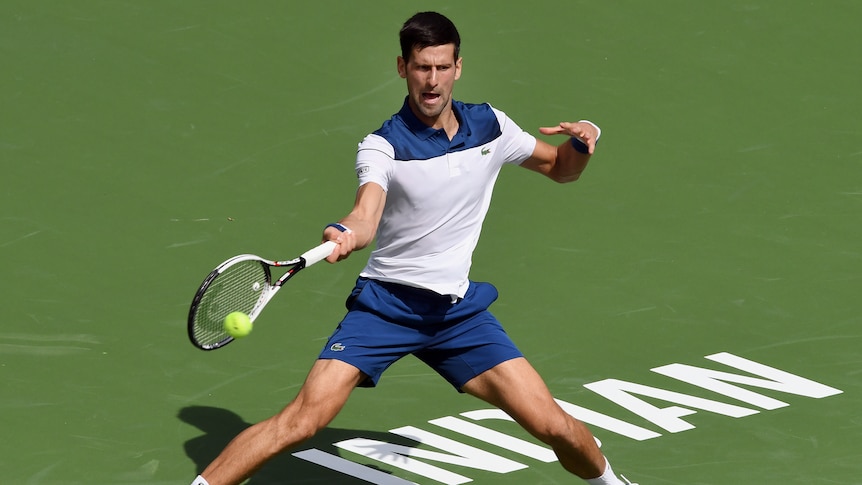 Novak Djokovic stretches to hit a forehand at Indian Wells, with the word "Indian" written in white on the court below h