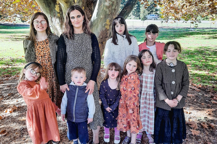 Mother with her nine children smile at the camera next to a tree.