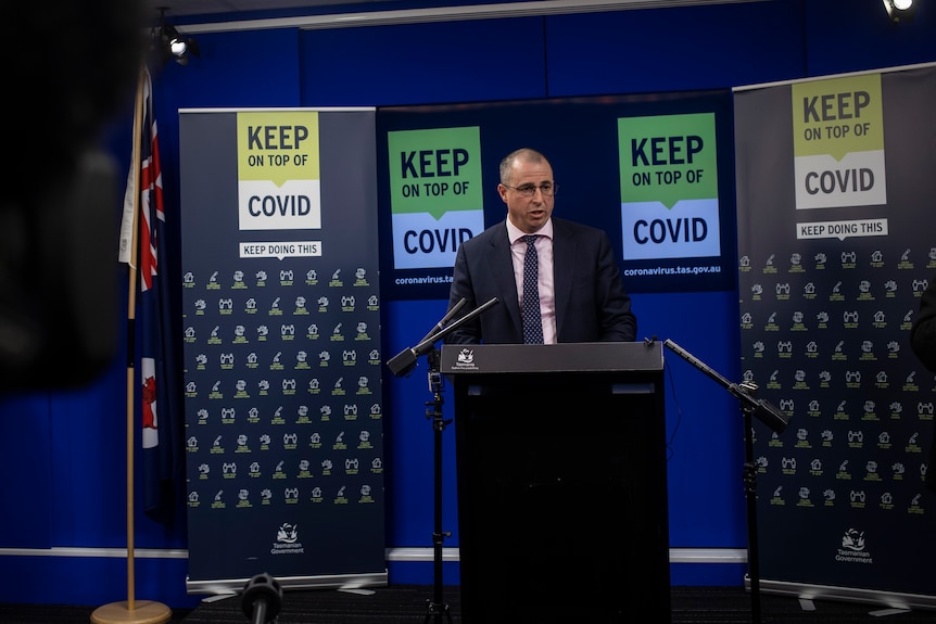 A man stands at a lectern in front of "keep on top of covid" banners and a Tasmanian flag
