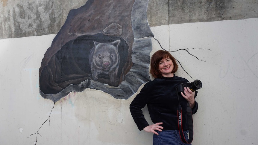 Bissland holding camera smiling and leaning against wombat mural.