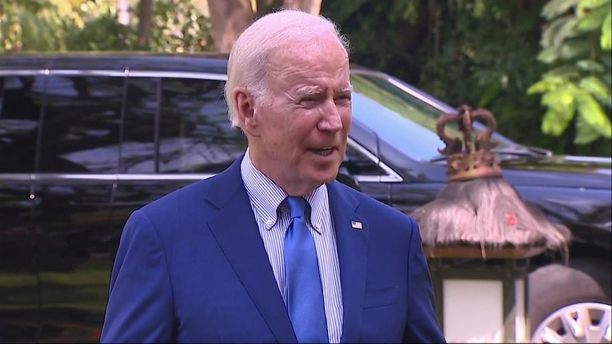 Joe Biden standing in front of a black vehicle, squinting, as he faces away to his left.