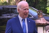 Joe Biden standing in front of a black vehicle, squinting, as he faces away to his left.