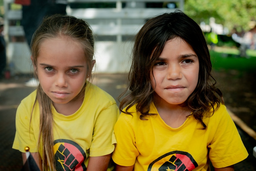 Two young girls sit on a bench in a park during an Invasion Day rally.