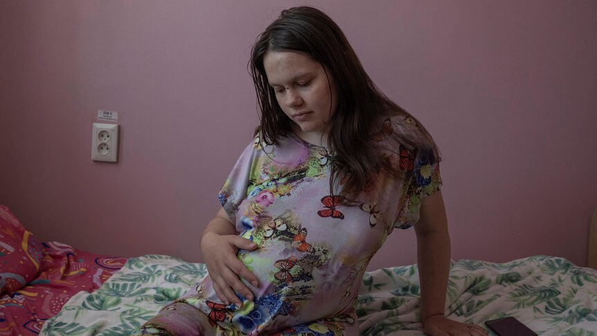 A teenage girl sitting on a hospital bed looks down at her pregnant belly where her hand rests. 
