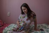 A teenage girl sitting on a hospital bed looks down at her pregnant belly where her hand rests. 