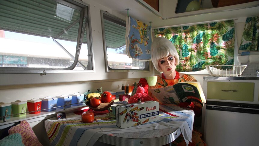 A man dressed in drag looks at a BP magazine at the table inside the caravan surrounded by 1960s decor