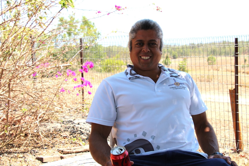 A man with white hair smiles at the camera, sitting on a chair, with landscape behind