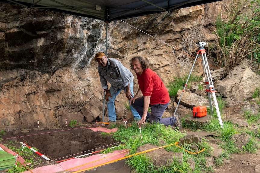 Two men lean over an excavated pit marked by string, with a gazebo roof above them.