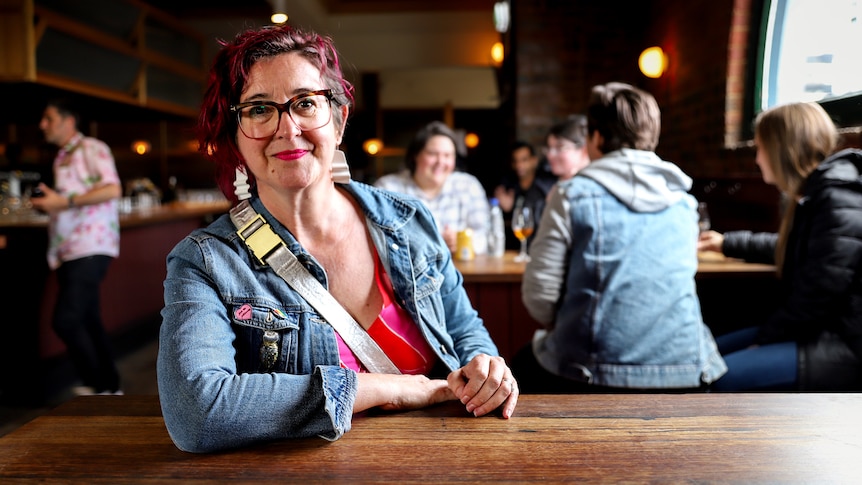 Woman with died pink hair and glasses wearing a denim jacket sits at a wooden table in a pub with other patrons in background
