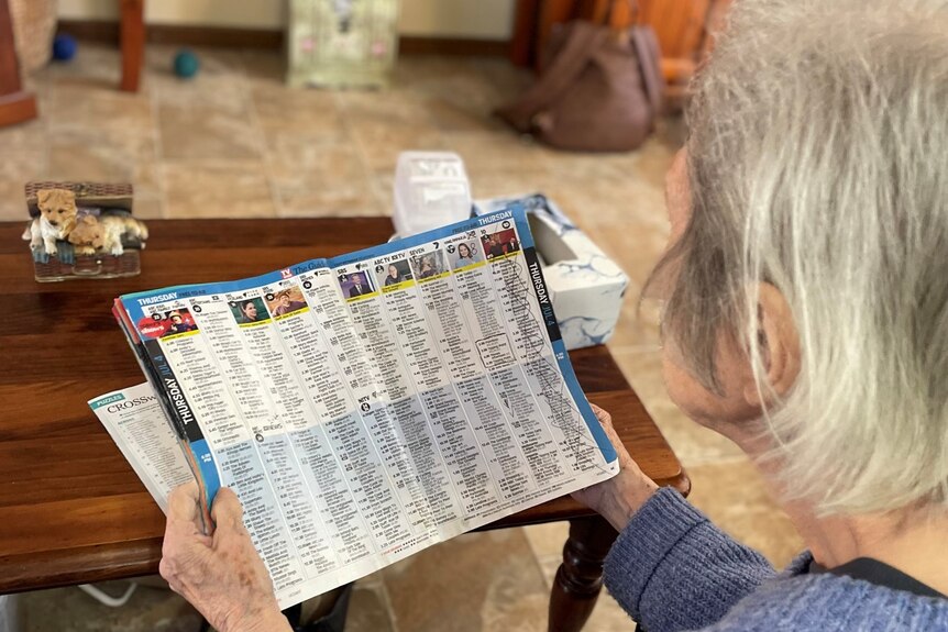 An elderly woman looking down at her television guide, with a coffee table in the background and