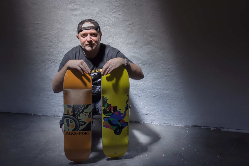 A man sits holding two colourfully designed skateboard decks