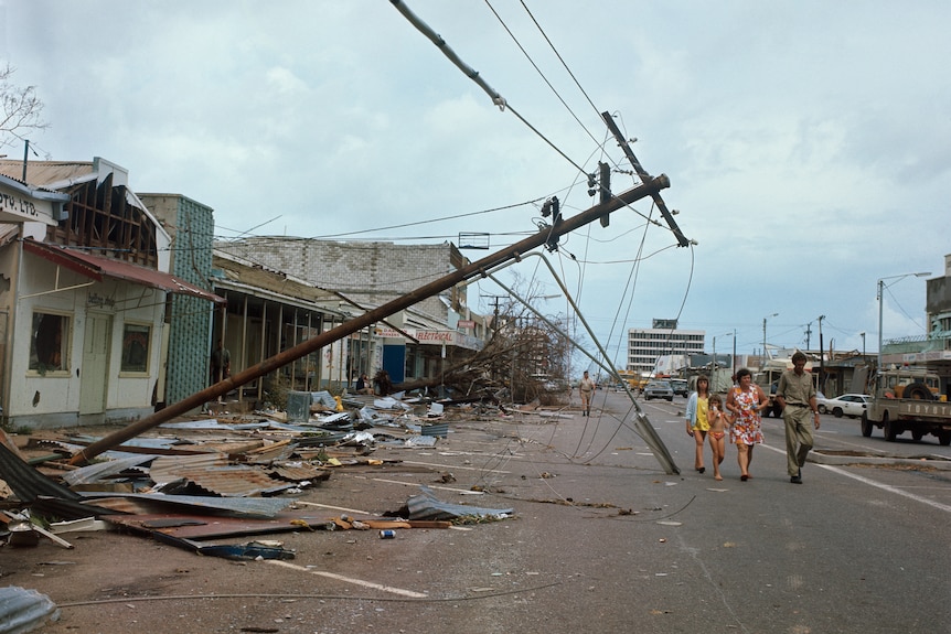 A family walks through a debris-laden main street. Shopfronts have awnings torn off. A broken power pole lies at an angle.